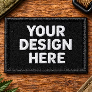 May include: A rectangular black patch with a stitched border featuring the white embroidered text YOUR DESIGN HERE in a bold, sans-serif font. The patch has a textured surface and is designed for attachment to gear or clothing items.