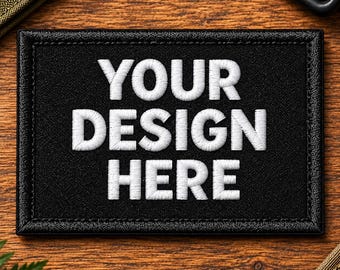 Puede incluir: Un parche rectangular negro con un borde cosido que presenta el texto bordado en blanco YOUR DESIGN HERE en una fuente sans-serif en negrita. El parche tiene una superficie texturizada y está diseñado para una personalización clara.