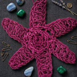 May include: A magenta crochet star with six points, alongside gemstones, safety pins, and dried lavender. The star is made with a textured stitch. Gemstones are blue and green. The star is a decorative item, possibly a coaster or embellishment.