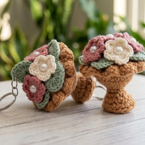 Op de afbeelding: Twee gehaakte paddenstoel sleutelhangers met lichtbruine stelen en hoeden, versierd met gehaakte bloemen in oudroze en crème. Het midden van elke bloem is geaccentueerd met een kleine witte parel, met groene gehaakte blaadjes tussen de bloemen.