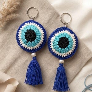May include: Two circular crochet evil eye keychains featuring concentric rings of black, light blue, white, and dark blue yarn. Each keychain includes a silver metal ring, a white bead, and a dark blue yarn tassel. Text reads EVIL EYE CROCHET PDF PATTERN.