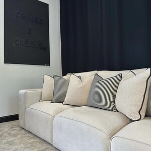 May include: A cream-colored sectional sofa with several decorative pillows. The pillows include solid cream-colored pillows, striped pillows, and pillows with black trim. A black wall art piece with the words "CROOKS & QUEENS" is visible.