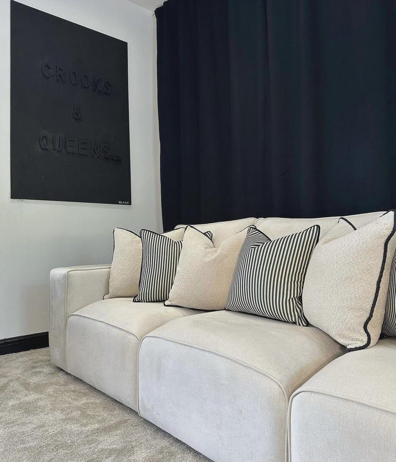May include: A cream-colored sectional sofa with several decorative pillows. The pillows include solid cream-colored pillows, striped pillows, and pillows with black trim. A black wall art piece with the words "CROOKS & QUEENS" is visible.