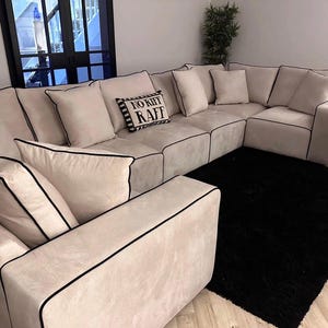 May include: A large, light-coloured sectional sofa with black piping. The sofa has several cushions and pillows. A black rug is in front of the sofa. A cushion with the text "NO RIFF RAFF" is on the sofa.