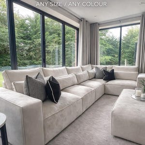 May include: A large, modular corner sofa in a neutral tone, with several cushions. The sofa is in a modern living room with large windows. Text on the image reads "MODULAR CORNER SOFA ANY SIZE / ANY COLOUR" and "Handmade in UK".