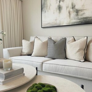 May include: A modern living room featuring a white sofa with decorative pillows. The pillows include solid cream-colored and black and white striped designs. Two round coffee tables with books and a bowl of green moss are in front of the sofa. A large abstract painting hangs on the wall.