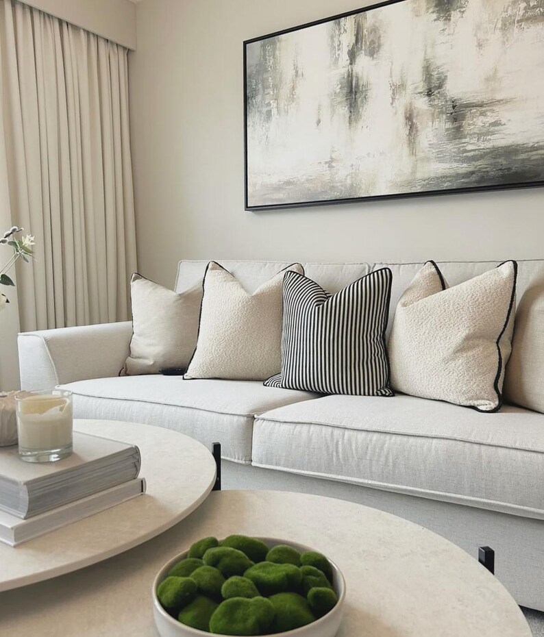 May include: A modern living room featuring a white sofa with decorative pillows. The pillows include solid cream-colored and black and white striped designs. Two round coffee tables with books and a bowl of green moss are in front of the sofa. A large abstract painting hangs on the wall.