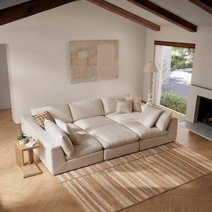 May include: A large, modular beige sofa with multiple cushions and pillows. The sofa sits on a striped rug in a living room setting. A small wooden side table with a plant is next to the sofa.