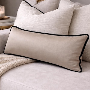 May include: A beige rectangular cushion with a black trim sits on a light-coloured sofa, with two other cushions. A cream-coloured knitted throw and a small side table with a candle are also visible.