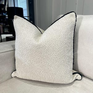 May include: A textured, off-white decorative pillow with a dark navy blue trim. The pillow is square-shaped and sits on a light-colored sofa. The pillow has a soft, plush appearance.