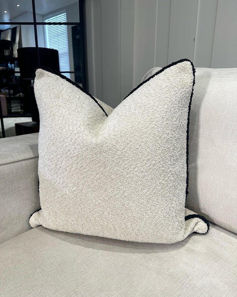 May include: A textured, off-white decorative pillow with a dark navy blue trim. The pillow is square-shaped and sits on a light-colored sofa. The pillow has a soft, plush appearance.