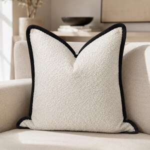 May include: A square, off-white decorative pillow with a textured surface and a black border. The pillow is placed on a neutral-toned sofa, suggesting a focus on home decor and interior design.