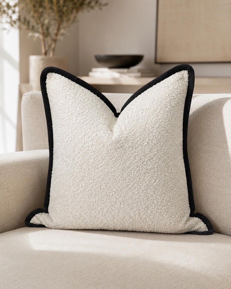 May include: A square, off-white decorative pillow with a textured surface and a black border. The pillow is placed on a neutral-toned sofa, suggesting a focus on home decor and interior design.