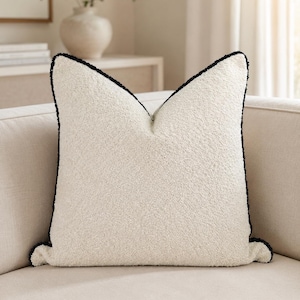 May include: A square, off-white decorative pillow with a textured surface and a black trim. The pillow is placed on a beige sofa, with a neutral-toned background. The pillow has a soft, inviting appearance.