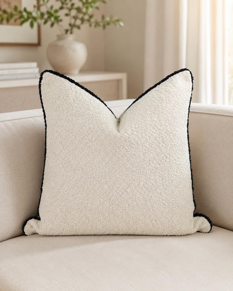 May include: A square, off-white decorative pillow with a textured surface and a black trim. The pillow is placed on a beige sofa, with a neutral-toned background. The pillow has a soft, inviting appearance.