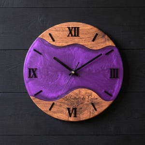 Puede incluir: Un reloj de pared redondo con secciones superior e inferior de madera natural separadas por un centro de resina púrpura con patrones de remolinos. La esfera muestra números romanos negros, manecillas negras y el texto grabado Heather & Galen.