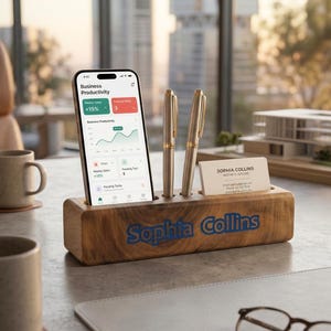 May include: A wooden desk organiser with the name "Sophia Collins" in blue lettering. It holds a smartphone displaying a business productivity app, two pens, and a business card. Two coffee mugs and a model building are also visible.