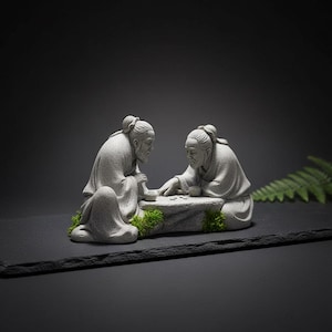 Pode incluir: Uma escultura em pedra cinzenta retrata duas figuras idosas absortas num jogo de tabuleiro. As figuras estão sentadas, com uma delas inclinada para a frente para colocar uma peça no tabuleiro. A escultura está sobre uma superfície escura com apontamentos de musgo verde.