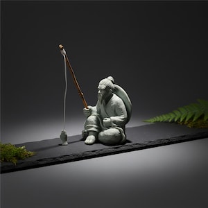 Peut inclure: Une sculpture en pierre grise d'un pêcheur assis, tenant une canne à pêche avec un leurre en forme de poisson. La figurine porte des vêtements traditionnels et a une longue barbe. La sculpture est posée sur une base rectangulaire noire.