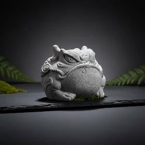 Puede incluir: Una escultura de rana de piedra gris con tallas detalladas, colocada sobre una superficie negra. La rana tiene una expresión intensa y está rodeada de musgo verde y hojas de helecho. La escultura es un objeto decorativo.