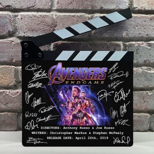 Può includere: Una clapperboard cinematografica nera con il titolo del film Avengers: Endgame e le firme del cast. La clapperboard ha una parte superiore a strisce bianche e nere e include la data di uscita: 26 aprile 2019.