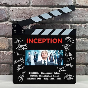 May include: A black movie clapperboard with the title "INCEPTION" in red. The clapperboard features a movie still, signatures, and text indicating the director, writer, and release date of July 13th, 2010.