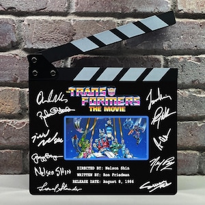 Puede incluir: Una claqueta de cine negra con el título "The Transformers: The Movie" y firmas. La claqueta incluye una imagen colorida de personajes de Transformers y la fecha de lanzamiento: 8 de agosto de 1986.