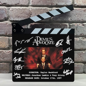 May include: A black film clapperboard featuring the title Devil's Advocate, a central image of two men in suits, and multiple white ink signatures. Text includes Director Taylor Hackford, writers Jonathan Lemkin and Tony Gilroy, and release date October 17th, 1997.