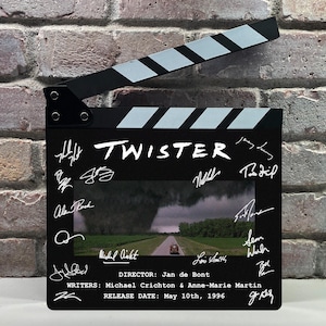 May include: A black film clapperboard featuring the title TWISTER in white text, a central image of a tornado over a road, and multiple white signatures. Text includes DIRECTOR: Jan de Bont, WRITERS: Michael Crichton & Anne-Marie Martin, and RELEASE DATE: May 10th, 1996.