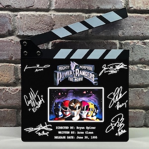 May include: A black movie clapperboard featuring the "Mighty Morphin Power Rangers: The Movie" logo and signatures. The clapperboard has a black and white striped top and a movie poster image.
