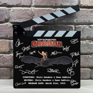 May include: A black movie clapperboard featuring the "How to Train Your Dragon" logo and signatures. The clapper has a black and white striped top and includes the release date: March 21st, 2010. The directors and writers are Chris Sanders & Dean DeBlois.