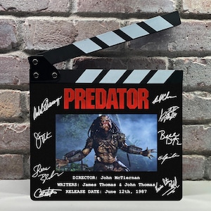 Puede incluir: Una claqueta de cine negra con la palabra "PREDATOR" en rojo. La claqueta incluye una foto del personaje Predator y firmas. La fecha de lanzamiento es el 12 de junio de 1987.