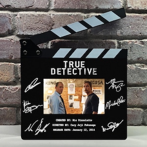 Puede incluir: Una claqueta de cine negra con el logotipo de True Detective, una escena central con dos personajes y varias firmas blancas. El texto incluye Created by Nic Pizzolatto, Directed by Cary Joji Fukunaga y Release Date January 12, 2014.