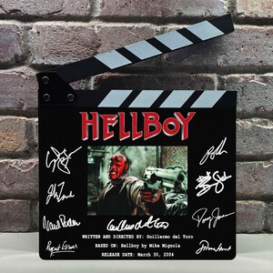 May include: A black film clapperboard featuring the title HELLBOY in red letters, a central image of the character Hellboy, and multiple white signatures. Text includes WRITTEN AND DIRECTED BY: Guillermo del Toro, BASED ON: Hellboy by Mike Mignola, and RELEASE DATE: March 30, 2004.