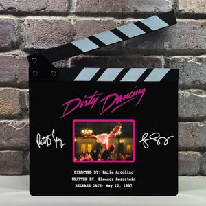 May include: A black movie clapperboard featuring the title "Dirty Dancing" in pink script. It includes a photo from the film, signatures, and the release date: May 12, 1987. The top has white and black stripes.