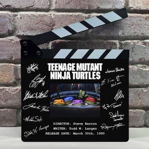 May include: A black movie clapperboard featuring the Teenage Mutant Ninja Turtles logo and signatures. The clapperboard has a black and white striped top and includes the director, writer, and release date.