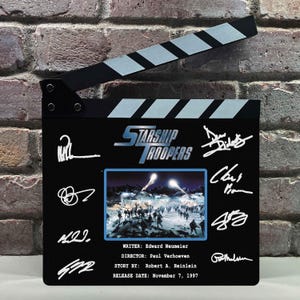 May include: A black movie clapperboard with the title "Starship Troopers" in blue and white. The clapperboard features a movie still, signatures, and text including the writer, director, story by, and release date: November 7, 1997.