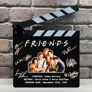 Könnte beinhalten: Eine schwarze Filmklappe mit dem "FRIENDS"-Logo in bunten Buchstaben. Die Klappe enthält ein Foto der Besetzung, Unterschriften und den Text "REGISSEUR: James Burrows" und "VERÖFFENTLICHUNGSDATUM: 22. September 1994."