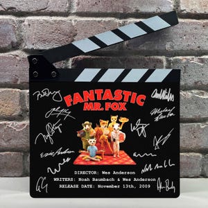 Puede incluir: Una claqueta de cine negra con el título "Fantastic Mr. Fox" en rojo y blanco, firmas y personajes de zorros. La claqueta también incluye al director, los guionistas y la fecha de lanzamiento.