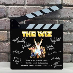 Peut inclure: Un clap de cinéma noir avec le titre "THE WIZ" en lettres dorées. Le clap présente des signatures et une image d'affiche de film. Le réalisateur, le scénariste et la date de sortie sont également indiqués.