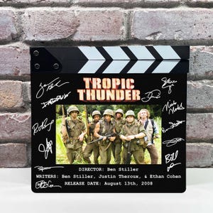 May include: A black movie clapperboard featuring the movie title "Tropic Thunder" in orange and white. The clapperboard has a photo of the cast in military attire. The director, writers, and release date are listed below the photo, along with several signatures.