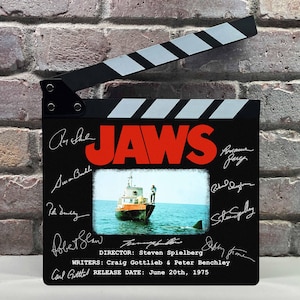 Op de afbeelding: Een zwarte filmklapper met het woord "JAWS" in het rood, handtekeningen en een filmstill van een boot en een haai. De klapper heeft een zwart-wit gestreepte bovenkant en bevat de regisseur, schrijvers en releasedatum.