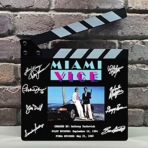 Op de afbeelding: Een zwarte filmklapper met het Miami Vice logo in lichtblauw en roze, een centrale foto van twee personen bij een zwarte auto en witte gedrukte handtekeningen. De tekst bevat CREATED BY: ANTHONY YERKOVICH, PILOT EPISODE: 16 september 1984, FINAL EPISODE: 21 mei 1989.