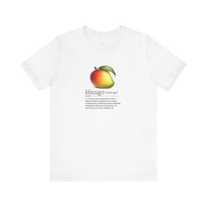以下が含まれることがあります： 白いTシャツ。マンゴーのリアルなイラストと定義が描かれています。マンゴーは赤、黄、緑の色合いで描かれ、緑の葉が付いています。下のテキストは「マンゴー」という言葉を定義しています。Tシャツは柔らかい素材で作られています。