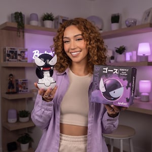 Può includere: Una statuetta di Gastly in bianco e nero che emerge da una base a forma di sfera bianca e nera, tenuta accanto alla sua scatola quadrata. La confezione riporta il testo Gastly, Ghost, buryle gas e caratteri giapponesi su un design viola e nero.