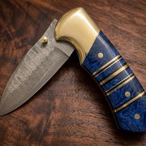 May include: A folding pocket knife featuring a Damascus steel blade with a wavy pattern, a polished brass bolster, and a handle crafted from blue marbled material accented with brass spacers and three circular brass pins along the grip.