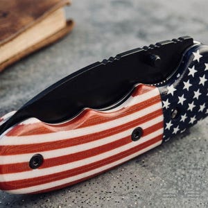 May include: A folding pocket knife with a black blade and a handle featuring the American flag design. The handle has red and white stripes on one side and a blue field with white stars on the other. The knife is closed and resting on a gray surface.