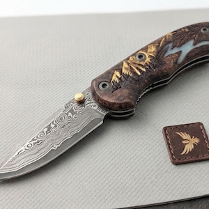 May include: A folding pocket knife with a Damascus steel blade featuring a wavy pattern and a gold-colored thumb stud. The handle is made of dark wood with an intricate carved mountain landscape design and a small square brown leather patch.