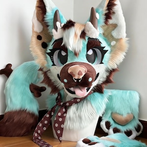 Può includere: Una testa e zampe di fursuit colorate nei toni del turchese, marrone e crema. La testa presenta grandi occhi espressivi, un naso rosa e una sciarpa marrone a pois bianchi. Sono visibili anche le zampe e la coda.