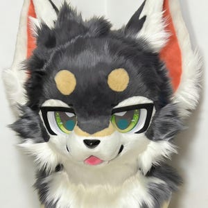 May include: A furry head with a wolf-like design. The head is primarily gray, with white and black accents. The ears have orange and white details. The eyes are green with black outlines, and the nose is black. The mouth is open, revealing a pink tongue.